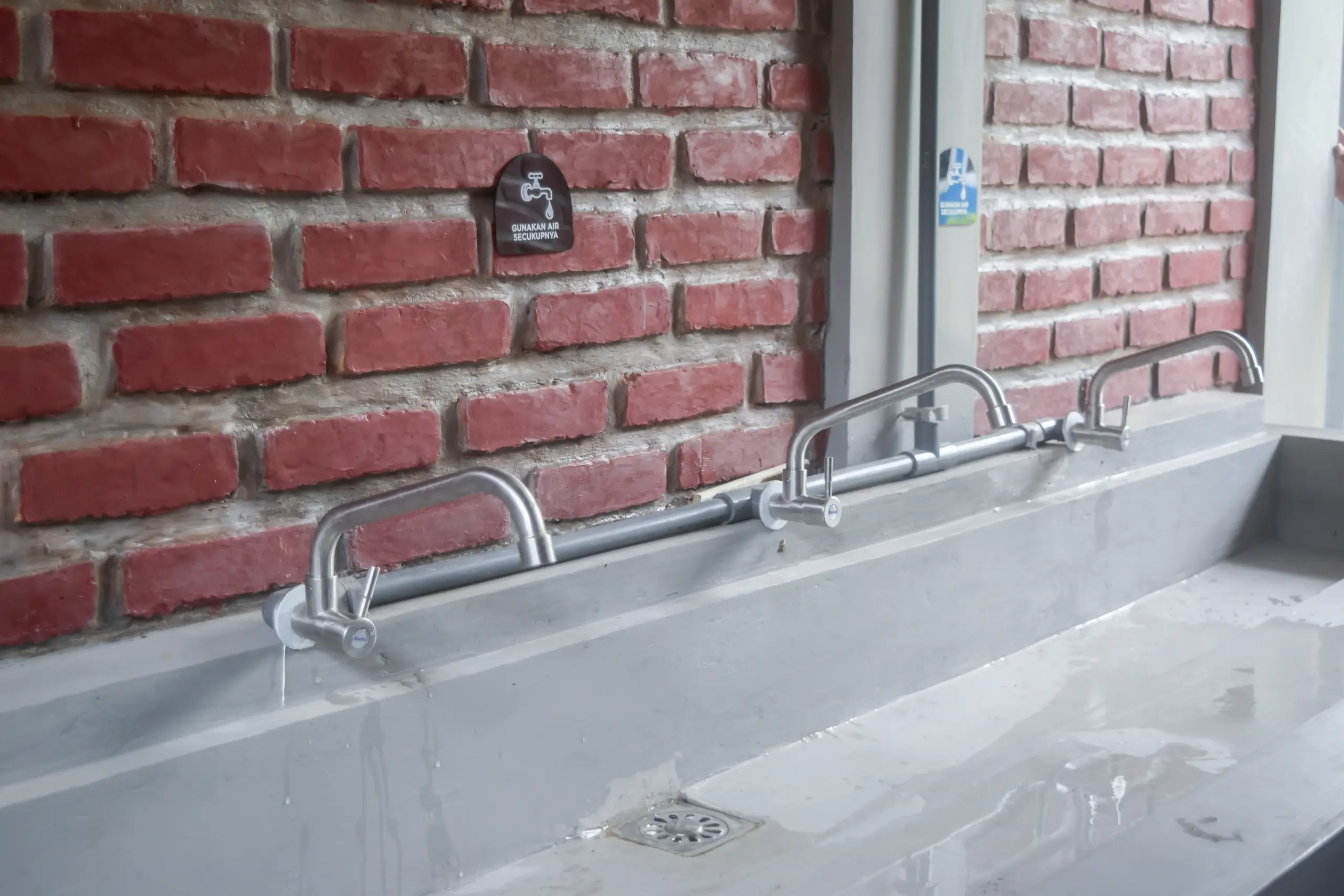 A concrete sink with steel faucets and a red brick wall behind, featuring a sign indicating water usage instructions.