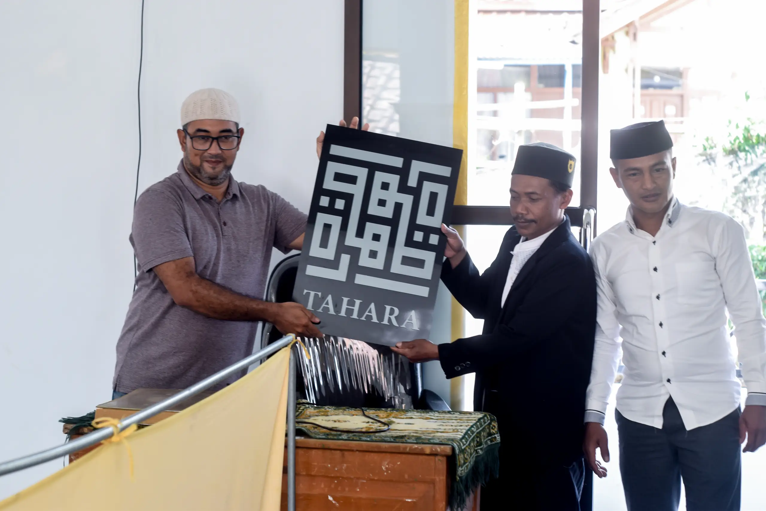 Two men hold a large sign reading "TAHARA," while a third man stands beside them in a decorated indoor space.