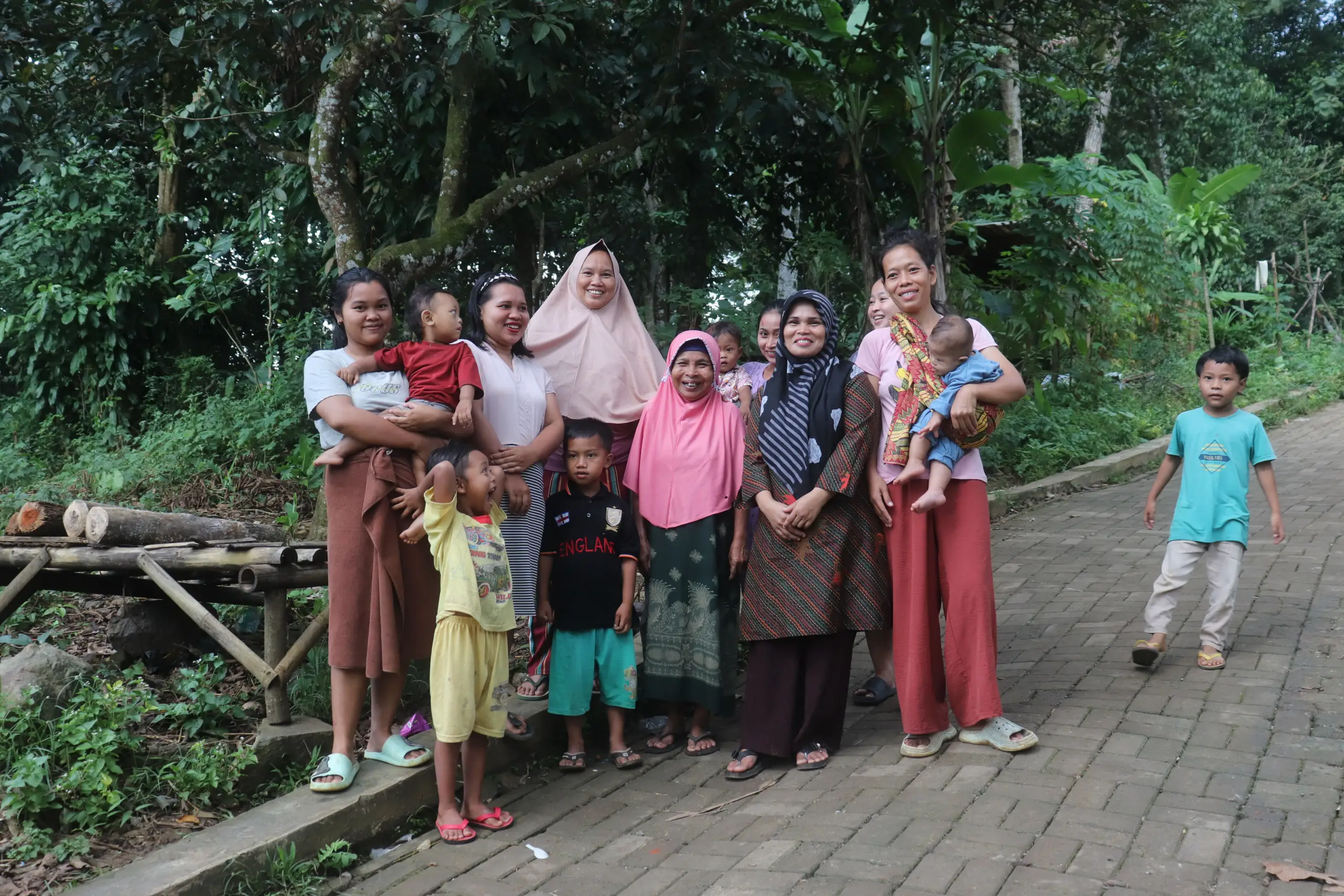 A group of women and children standing together outdoors among lush greenery, with a pathway and logs visible in the background.