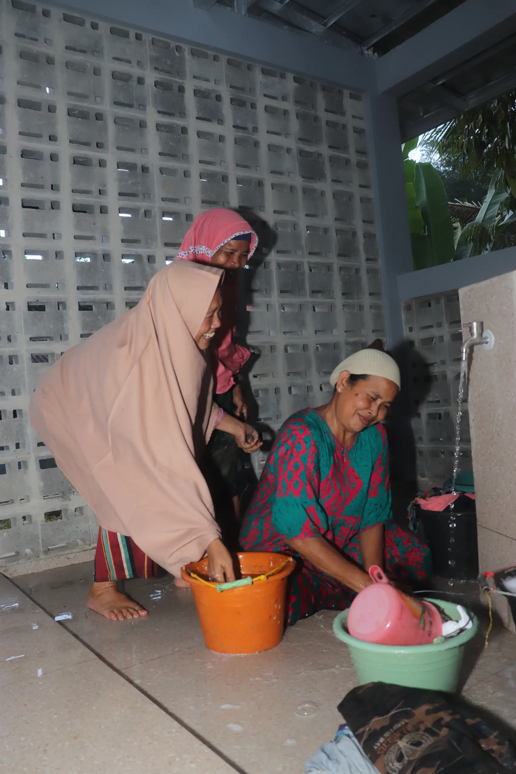Two women in traditional clothing wash clothes together in a covered outdoor space, with buckets and garments scattered around.