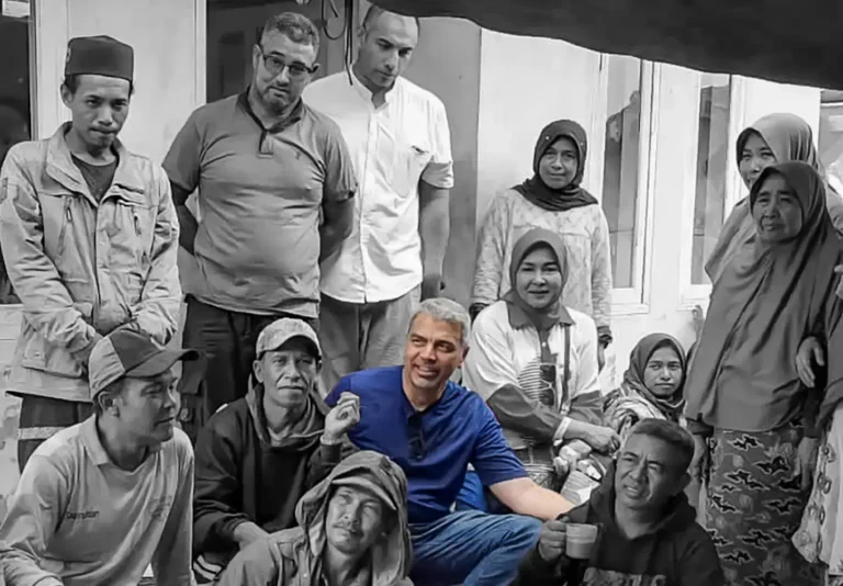 A diverse group of individuals gathered together, mostly seated, in a casual outdoor setting. They wear casual clothing and appear engaged.