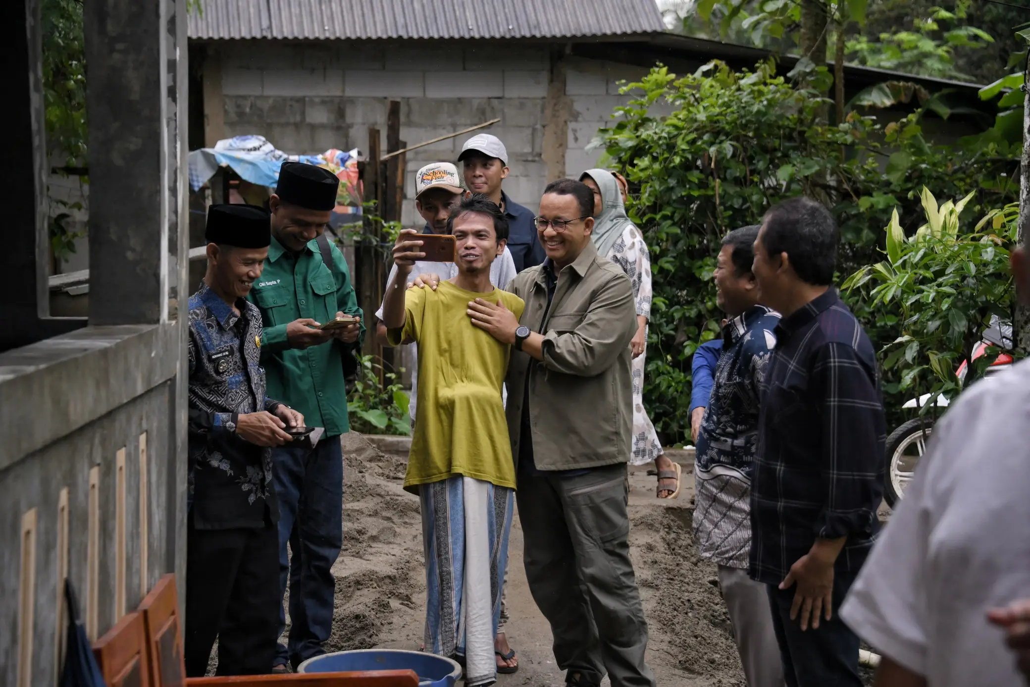 A group of people in casual clothing gather outdoors, engaged in conversation, with greenery and a partially constructed building in the background.