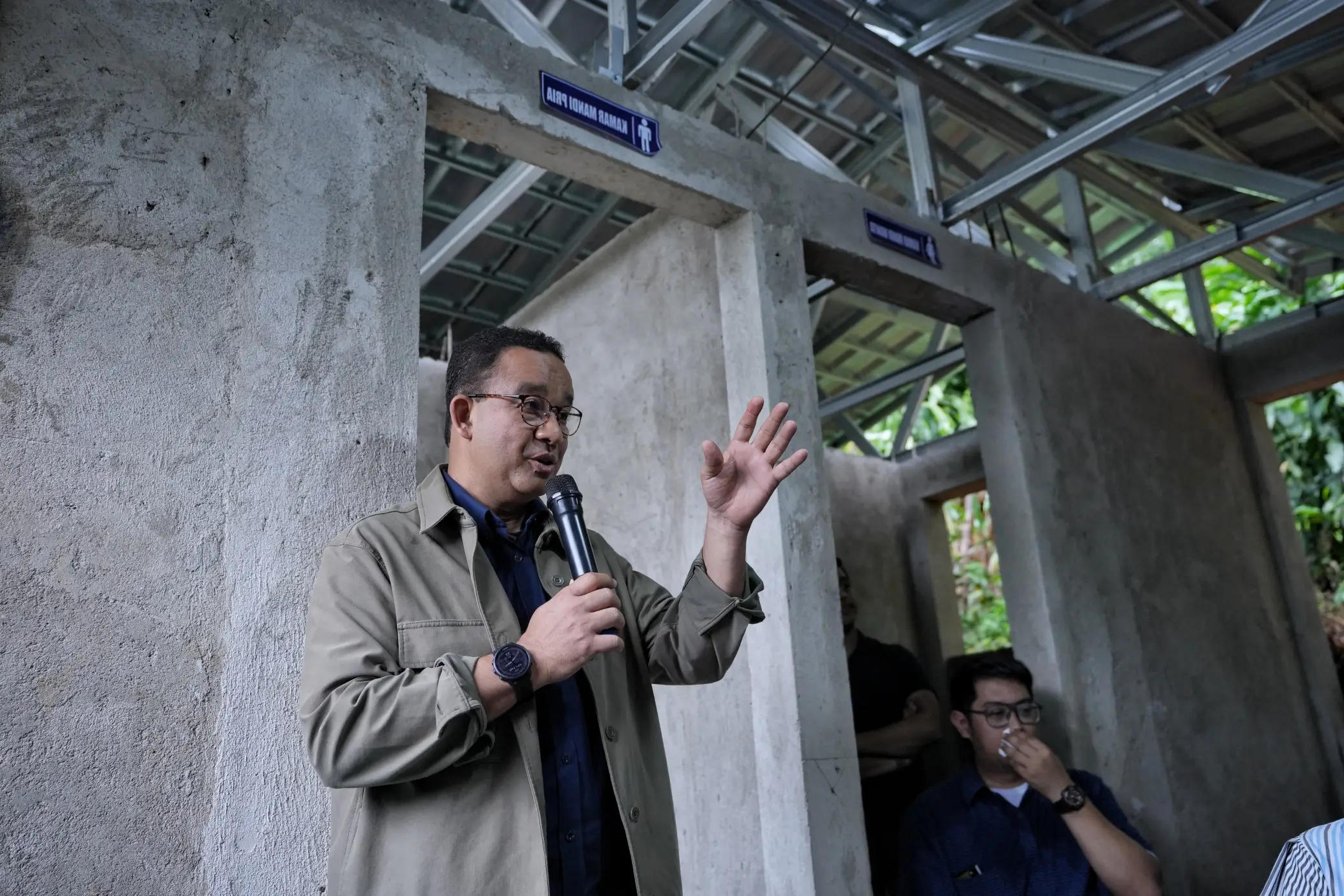 A person stands with a microphone, gesturing, in a partially constructed building with restroom signs above openings.