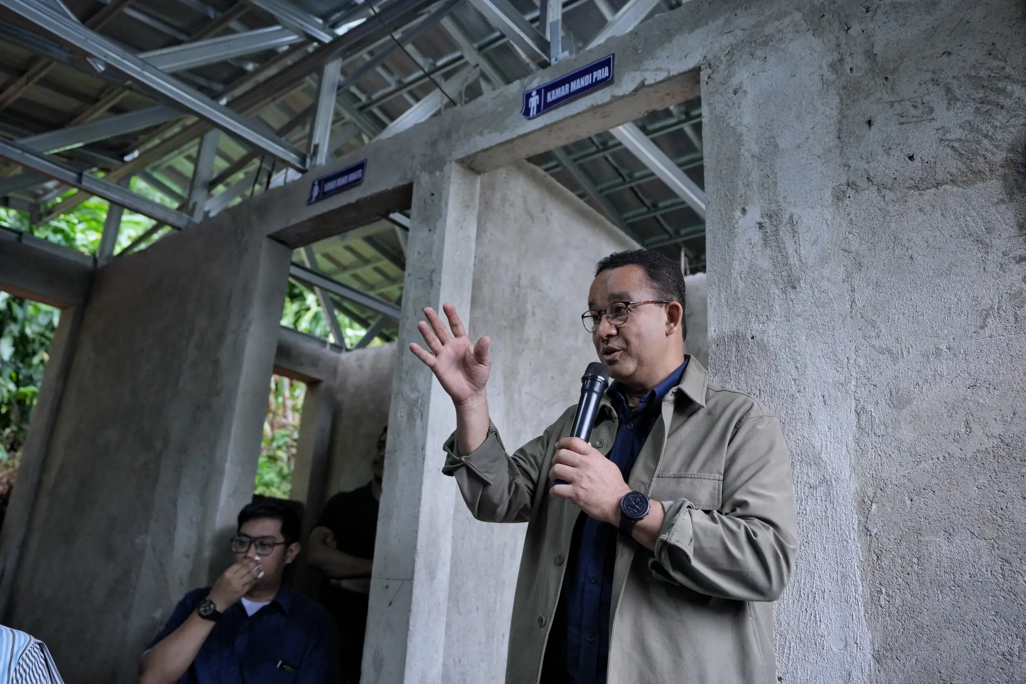A person stands with a microphone, gesturing, in a partially constructed building with restroom signs above openings.