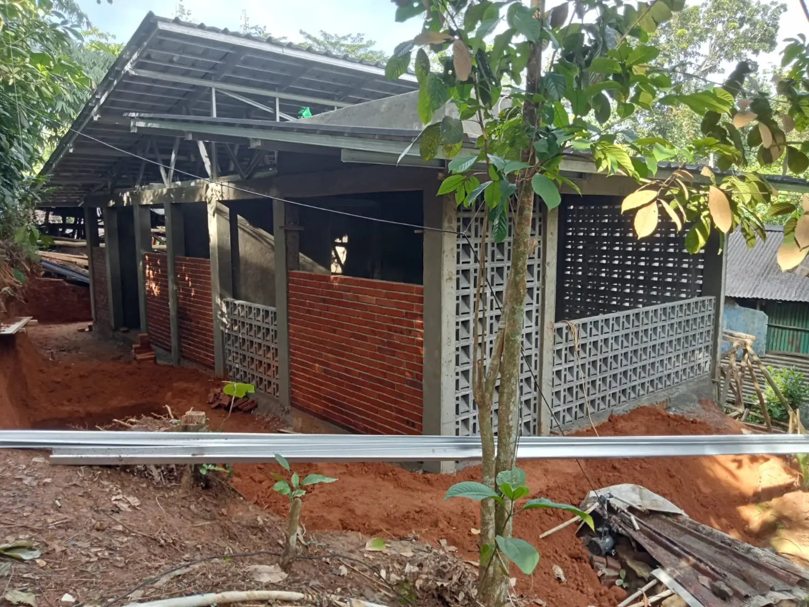 A partially constructed house with brick and concrete walls, a metal roof, surrounded by red soil and greenery in a rural setting.