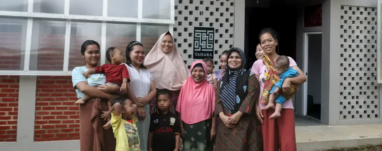 A group of women and children stand together in front of a building with a sign that reads "TAHARA,".